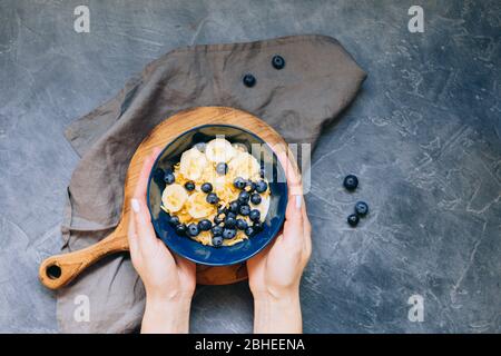 Frau Hände halten dunkelblaue Schale Haferbrei Haferbrei mit Banane und Heidelbeere auf Vintage-Tisch Top-Ansicht in flacher Lay-Stil. Warmes Frühstück und hausgemacht Stockfoto