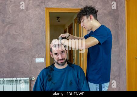 Bärtiger Mann, der zu Hause seine Haare schneidet, half seinem jungen Sohn mit lockigen Haaren im Wohnzimmer zu Hause Stockfoto
