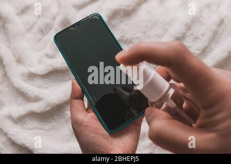 Menschliche Hand hält ein Antiseptikum und puffert Flüssigkeit auf dem Smartphone, um das Gerät zu reinigen. Stockfoto