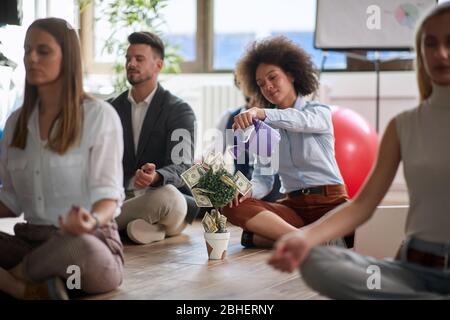 Geschäftskollegen meditieren bei der Arbeit, sitzen auf dem Boden. Afro-amerikanische Frau macht Visualisierung von Geld Einkommen erhöhen, Bewässerung Bonsai-Baum Stockfoto