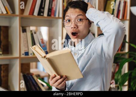 Bild von staunenden asiatischen Mann in Brillen zum Ausdruck Schock und Buch lesen, während in der Bibliothek stehen Stockfoto