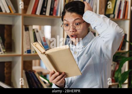 Bild von staunenden asiatischen Mann in Brillen zum Ausdruck Schock und Buch lesen, während in der Bibliothek stehen Stockfoto