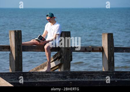 Portobello, Schottland, Großbritannien. 25. April 2020. Blick auf die Menschen im Freien am Samstagnachmittag am Strand und an der Promenade in Portobello, Edinburgh. Gutes Wetter hat mehr Menschen ins Freie gebracht, die wandern und Radfahren. Der Strand scheint voll mit möglicherweise einem Zusammenbruch der sozialen Distanz, die später am Nachmittag stattfindet. Einsamer Mann, der Buch liest, auf Holzgroyne sitzend. Iain Masterton/Alamy Live News Stockfoto