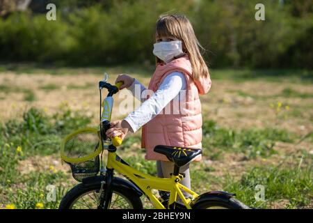 Ein kleines Mädchen, das an einem sonnigen Tag Gesichtsmaske auf einem Fahrrad trägt. Kinder, Spielzeit während der Epidemie. Corona Virenschutz für Kinder. Stockfoto