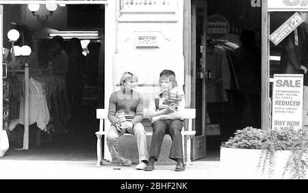 Das tägliche Leben auf den Straßen in Manchester, England, Großbritannien im Jahr 1974. Zwei junge Teenager sitzen auf einer Bank vor einem Kleiderladen.