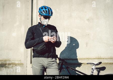 Der Kurier in Schutzmaske auf dem Fahrrad hielt an und beobachtete die Telefonanwendung. Lieferservice in Quarantäne. Ein Mann in einem Atemschutzgerät verwendet ein Stockfoto