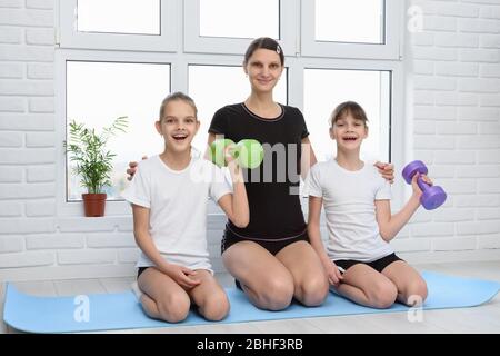 Zwei fröhliche Mädchen und ihre Mutter sitzen auf dem Boden mit Hanteln auf der Matte Stockfoto