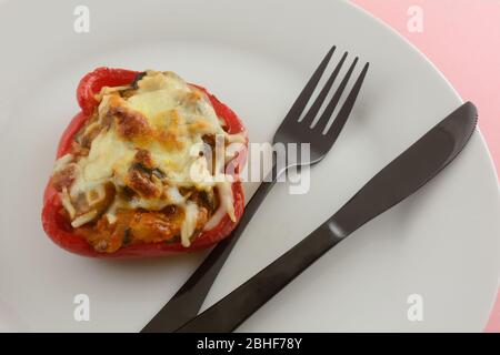 Gefüllte rote Paprika mit Käse auf weißem Teller mit schwarzer Gabel und Messer Stockfoto