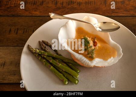 Meeresfrüchte-Mahlzeit von Hummer Biskuitsuppe in einer Muschelschale auf einem weißen Teller mit Spargel. Stockfoto