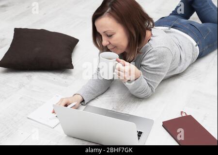 Schöne Frau mit Laptop liegt auf dem Boden im Wohnzimmer. Freiberuflicher Mitarbeiter, der zuhause arbeitet. Stockfoto
