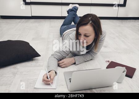 Schöne Frau mit Laptop liegt auf dem Boden im Wohnzimmer. Freiberuflicher Mitarbeiter, der zuhause arbeitet. Stockfoto