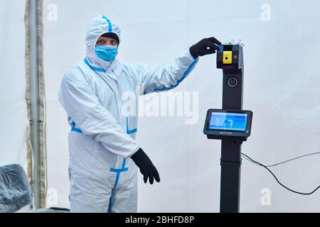 Perm, Russland - 25. April 2020: Der medizinische Offizier im Schutzanzug lädt zur berührungslosen Temperaturmessung im Zelt des Sanitär- und Sanitätsanzugs ein Stockfoto