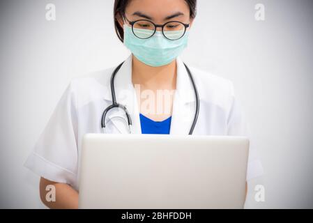 Studio Portrait Asiatische junge Frau Arzt trägt Brille und Maske zur Sicherheit Coronavirus Blick auf den Laptop, Arzt mit Stethoskop arbeiten an Stockfoto