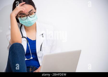 Asiatische junge Frau Arzt tragen Maske zur Sicherheit Coronavirus sitzen auf dem Boden Blick auf den Laptop mit Traurigkeit, Stress und Sorgen über das Problem, Conc Stockfoto