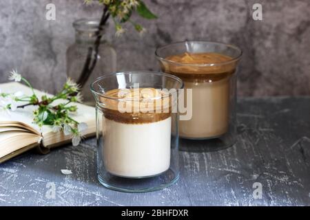 Dalgona Kaffee in transparenten Gläsern, ein Buch und Blumen auf einem Betontisch. Stockfoto
