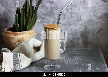 Dalgona Kaffee in einer transparenten Tasse mit einem Cocktail Stroh und Kaktus auf einem Betontisch. Stockfoto