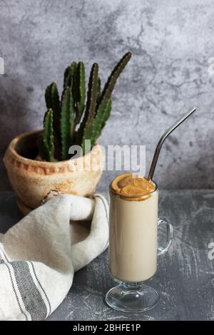Dalgona Kaffee in einer transparenten Tasse mit einem Cocktail Stroh und Kaktus auf einem Betontisch. Stockfoto