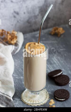 Dalgona Kaffee in einer transparenten Tasse mit einem Cocktail-Stroh, Kekse und getrockneten Blumen auf einem Betontisch. Stockfoto