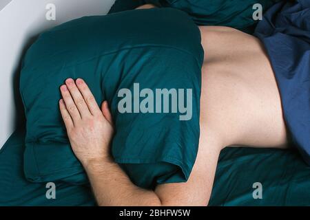 Ein schöner junger Mann, der sich Kopf und Ohren bedeckt und versucht, den Klang mit einem Kissen abzublocken, während er von lauten Geräuschen wach gehalten wird Stockfoto