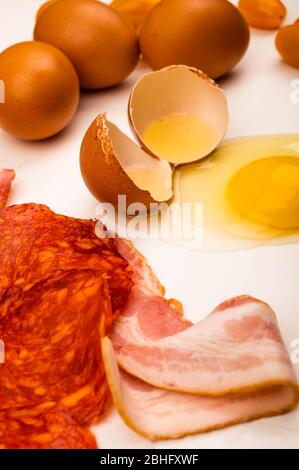 Zerbrochenes Hühnereier, verstreute Hühnereier, Scheiben Wurst und Speck und Tomaten auf weißem Hintergrund. Nahaufnahme Stockfoto