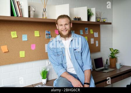 Lächelnder junger Erwachsener bärtiger Gelegenheitsmann, der Kamera im Heimbüro anschaut. Stockfoto
