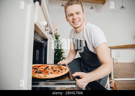 Der Küchenchef lächelt und nimmt Backblech mit heißen Pizzatomaten und Oliven, im Hintergrund helle Küche. Kochen Blogger Konzept Stockfoto