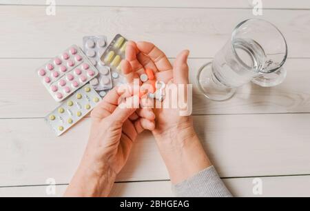 Ältere Hände mit Pillen und Drogen auf dem hellen Tisch, Glas Wasser. Faltige Hände der alten Frau mit bunten Tabletten, Holzhintergrund. Schmerzhaftes altes Stockfoto