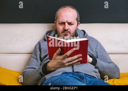 Ernst junger bärtiger Mann in lässiger Kleidung entspannt auf Couch Lesung rotes Buch auf schwarzem Hintergrund Stockfoto