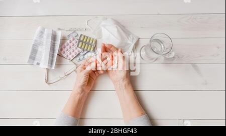 Ältere Hände, Pillen und Medikamente auf dem Tisch, Glas Wasser. Faltige Hände der alten Frau mit bunten Tabletten, Maske, Brille, Rezept, Thermometer Stockfoto