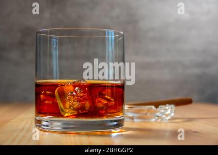 Aschenbecher mit einer Zigarre im Hintergrund in Unschärfe-Effekte. Glas Whiskey mit Eiswürfeln auf dem Holztisch Stockfoto