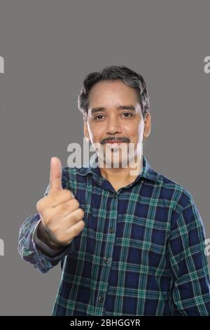 Lächelnder indischer Trader Mann mit Schnurrbart zeigt Thumbs Up auf grauem Hintergrund Stockfoto