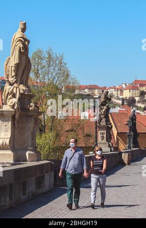 Prag, Tschechische Republik - 23. April 2020: Ehepaar mittleren Alters, die auf der Karlsbrücke mit medizinischen Gesichtsmasken spazieren. Statuen auf der Brücke. Stadtzentrum während der Coronavirus-Pandemie. COVID-19-Krise. Stockfoto