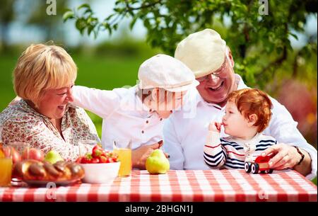 Glückliche Großeltern mit Enkelkindern sitzen am Schreibtisch im Frühlingsgarten Stockfoto