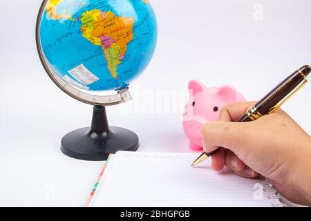 Lady Hand halten Stift mit leeren offenen Notizbuch mit Erdkugel und Sparschwein auf Schreibtisch. Stockfoto