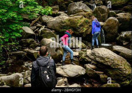 Gemischte Gruppe von Touristen auf Felsbrocken am Torc Waterfall im Killarney National Park, County Kerry, Irland Stockfoto