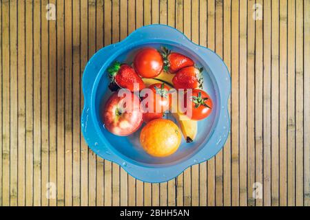 Blaue Schale mit verschiedenen frischen und sauberen Früchten auf Holzbasis. Bananen, Tomaten, Äpfel, Erdbeeren und Orangen im Wasser. Stockfoto