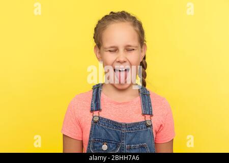 Porträt von frechen kleinen Mädchen in Denim Overalls herausstechen Zunge und Augen geschlossen, ungehorsam Kind herumtäuschen mit lustigen spöttischen expr Stockfoto