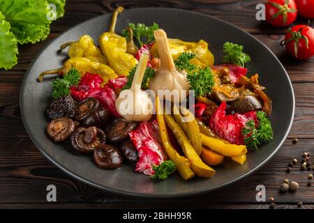 Gesalzenes und eingelegtes Gemüse: Gurken, Knoblauch, Pfeffer, Pilze, Kohl, Eierplanton ein schwarzer Teller auf einem hölzernen Hintergrund Stockfoto