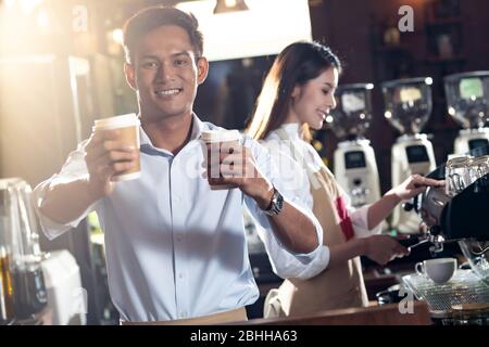 Porträt des asiatischen Barista, der dem Gast im Café eine Tasse Kaffee zum Mitnehmen serviert, während ein anderer Kollege im Hintergrund arbeitet. Für kleine Unternehmen Stockfoto