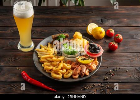 Ein Glas schäumenden Biers und ein Teller mit Snacks: Garnelen, Tintenfischringe, pommes Frites, Roggencroutons, Oliven auf einem hölzernen Hintergrund Stockfoto