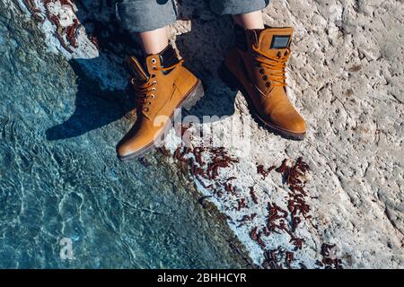 Unkenntlich Frau in gelben Trekking-Stiefel sitzt an der Küste in der Nähe von türkisfarbenem Wasser, Draufsicht. Abenteuer, Scout, Entdeckung, Entdecker Konzept Stockfoto