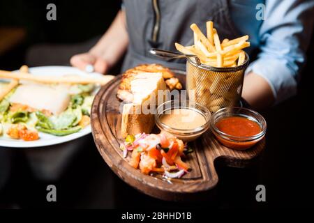 Korb mit pommes frites. Serviert mit gegrillter Soße und Flügeln auf einem Holzteller. Stockfoto