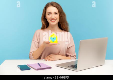 Versicherungsagentur. Fröhliche hübsche Frau, die Papierhaus mit Herz im Inneren hält und lächelt, Immobilienmakler, der am Schreibtisch mit Laptop sitzt, Heimbüro. Stockfoto