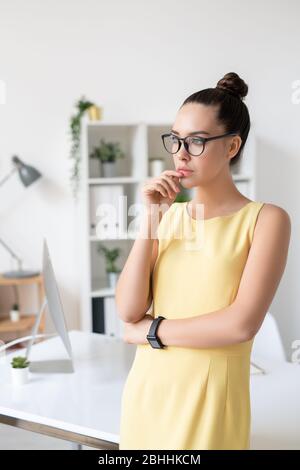 Nachdenkliche unternehmungslustige junge Geschäftsfrau in gelbem Kleid und Brille mit der Hand in der Nähe der Lippen im modernen Büro Stockfoto