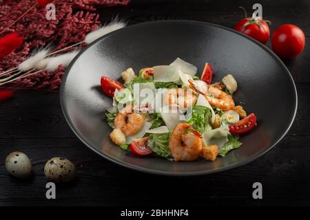 caesar Salat mit Garnelen in einem schwarzen Teller auf einem dunklen Holzhintergrund Stockfoto