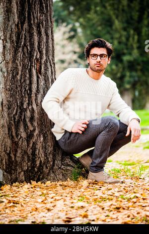 Schöner junger Mann allein in der Natur in der Nähe eines Baumes im Freien. Ein charmanter junger Mann mit Brille. Der schöne Junge hockt auf dem Boden lehnt die t Stockfoto