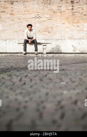 Junger Mann sitzt auf einer Marmorbank in der Straße in der Stadt. Ernsthafte Einstellung. Weißer Pullover. Ellbogen an den Beinen. Kopierbereich. Stockfoto