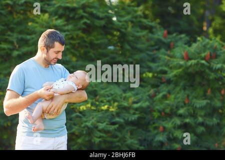 Vatertag. Der fürsorgliche Vater hält das Baby im Park auf einem grünen Blatthintergrund in den Armen. Stockfoto