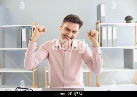 Positiv motivierter Geschäftsmann im Hemd hob seine Arme lächelnd an, während er am Tisch im Raum saß. Stockfoto