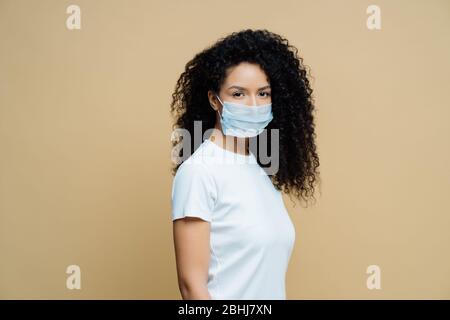 Seitliches Schuss von Afro American Frau trägt schützende Gesichtsmaske, schützt vor der Ausbreitung von Coronavirus-Krankheit, bleibt zu Hause, folgt Regeln der selbst Stockfoto